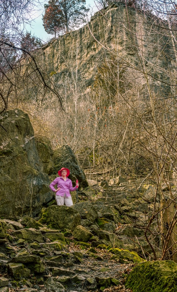 Rocks, trees, women in red hat and pink shirt and gray pants with walking stick.