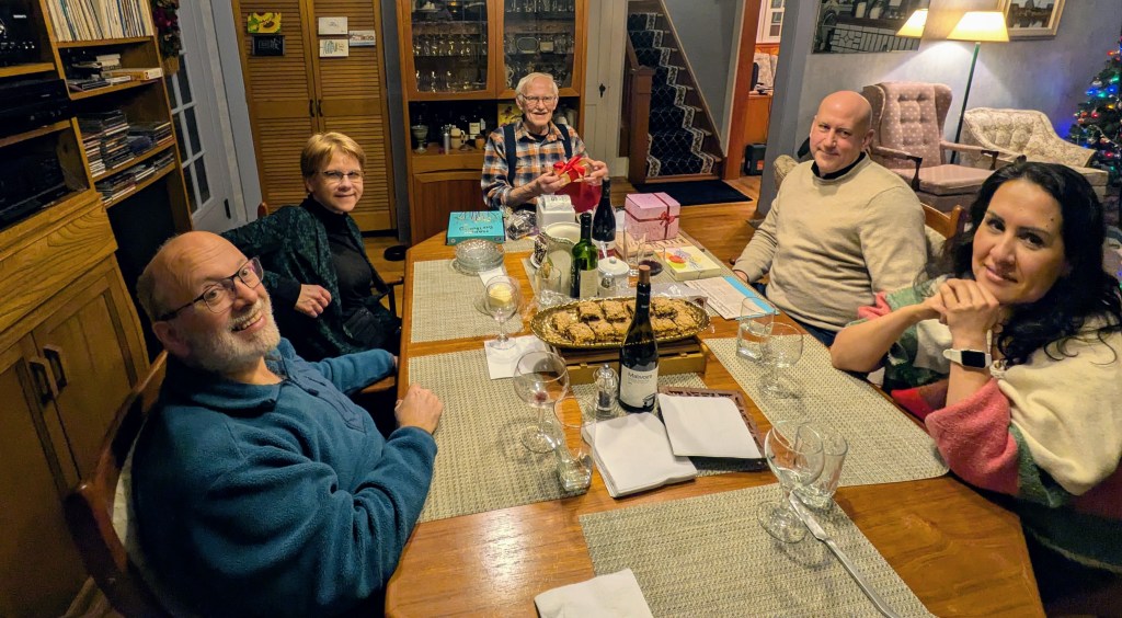 Table with five people, desserts, wine, and some birthday gifts. Christmas tree in the corner.