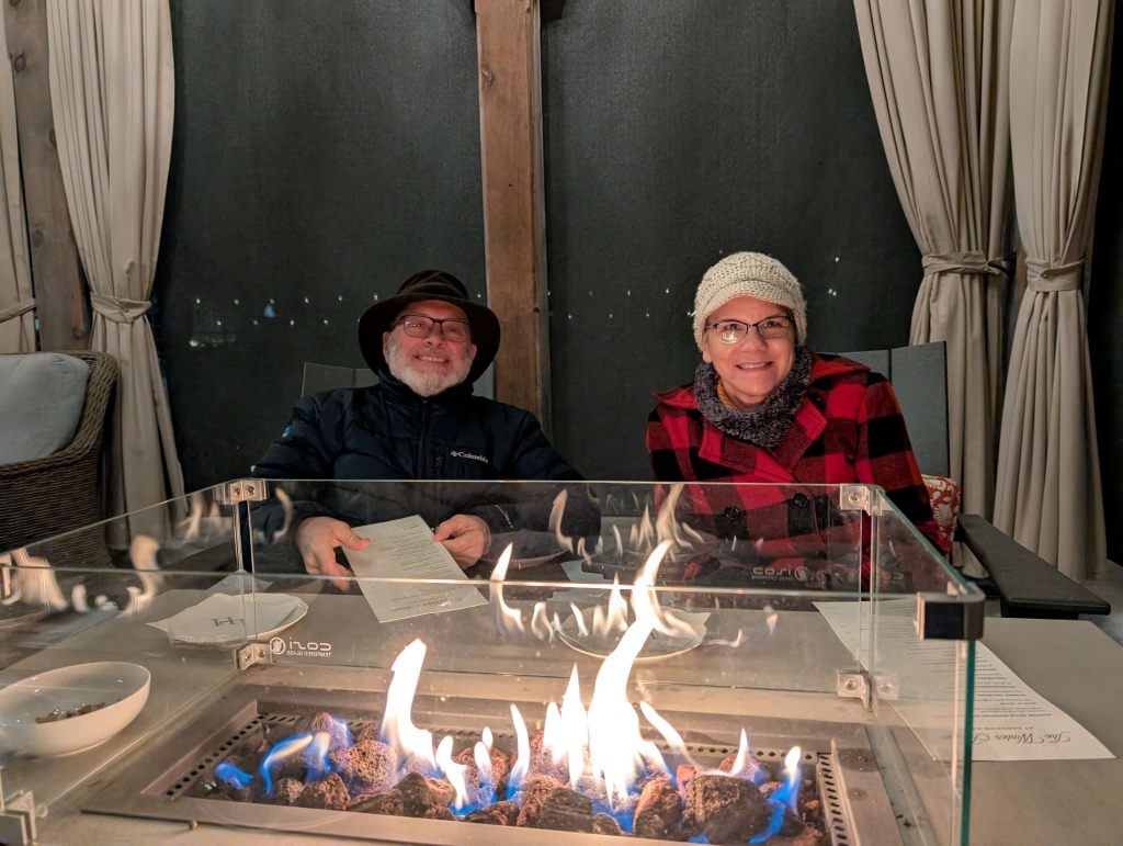 Couple in hats and coats at a table in front of a fire.