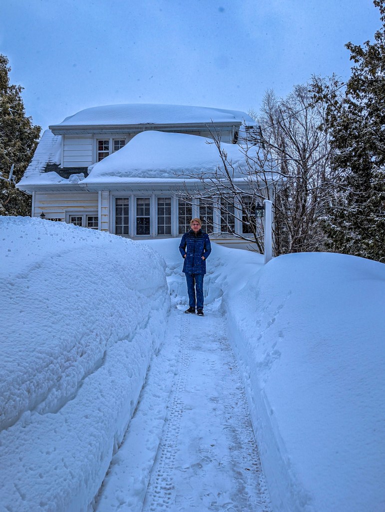 Woman in front of snow-covered house surrounding by very tall snow banks.