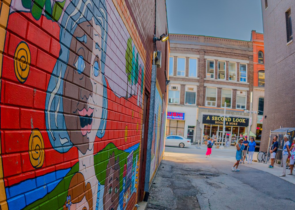 Side view of a mural showing a young girl, down an alley, with group to the side and A Second Look Books & More sign in the distance.