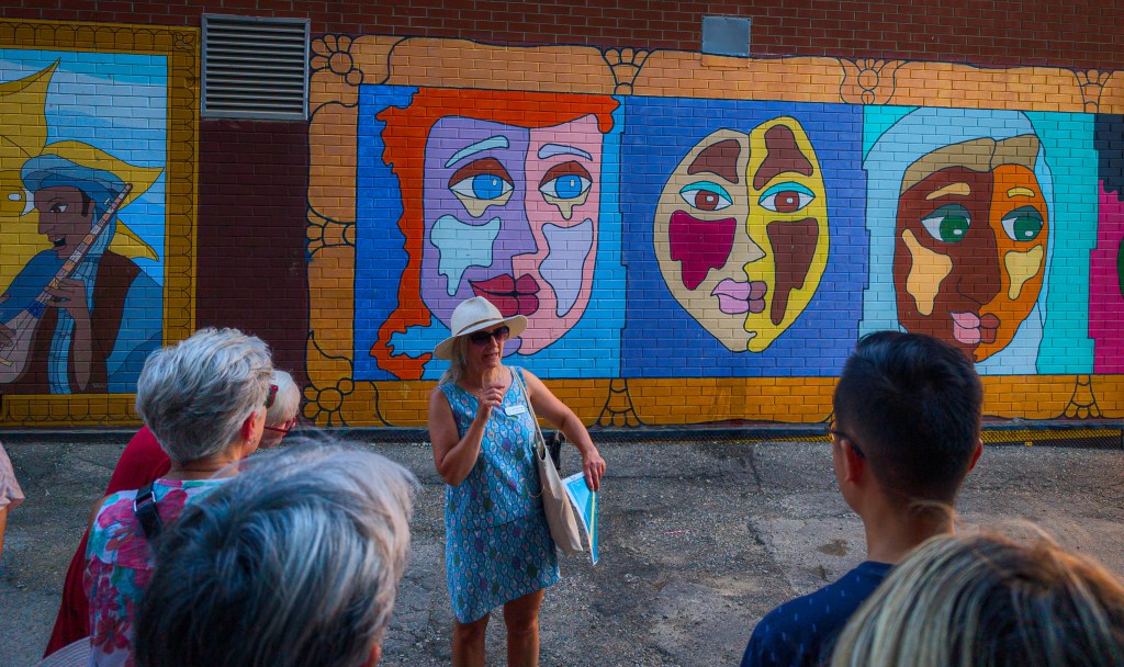Woman speaking to a group in front of a mural depicting three colourful faces.