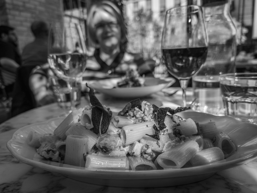 Black and white photo of a plate of pasta with two glasses of wine, and woman in the background.